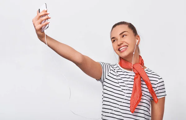 Studio horisontellt porträtt av vacker europeisk ung kvinna tar självporträtt över vit vägg, klädd randig t-shirt och röd halsduk på halsen. Lycklig kvinna gör en selfie på hennes smarta telefon. — Stockfoto