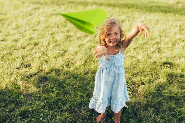 Boldog csinos kislány játszik a papír sík a nyári nap a parkban. Aranyos gyerek dobott egy repülőgép a szabadban a kertben. Gyermekkori koncepció — Stock Fotó