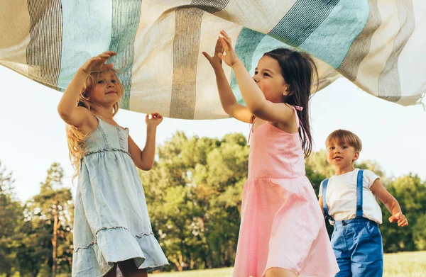 담요 아래에서 놀고, 점프하고, 함께 춤을 추는 아이들. 공원에서 여름 날을 즐기는 행복한 어린 소년과 어린 소녀. 야외에서 놀고있는 쾌활한 아이들. 어린 시절 개념 — 스톡 사진