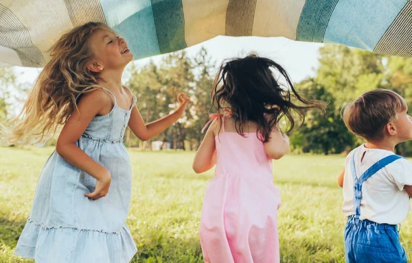 Crianças brincalhões brincando sob o cobertor, pulando e dançando juntos. Menino e garotinhas felizes curtindo o dia de verão no parque. Miúdos alegres a brincar ao ar livre. Conceito de infância — Fotografia de Stock