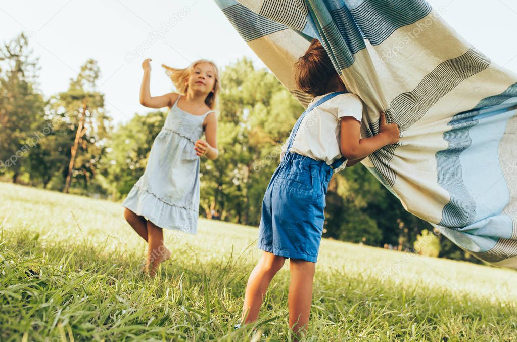 Outdoors image of children playing under the blanket, jumping and dancing together. Happy little boy and little girl enjoying summer day in the park. Cheerful kids playing outdoors. Childhood concept