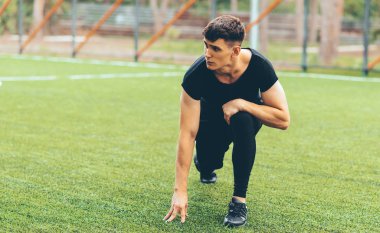 Sağlıklı genç adam egzersiz ve yeşil çim üzerinde streching. Atlet adam bir kenara bakıyor, bacağını esnetiyor ve kulaklıkla müzik dinliyor. Spor, yaşam tarzı ve insan kavramı.