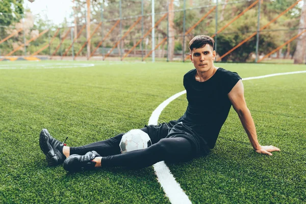 Bonito jovem jogador de futebol homem tendo um descanso após a sessão de treinamento ao ar livre. Homem saudável e apto sentado e relaxante no gramado verde após exercícios de treino. Pessoas e conceito de desporto — Fotografia de Stock