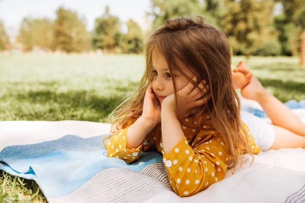 Nettes kleines Mädchen, das auf der Decke im grünen Gras liegt und es sich bequem macht. schönes Kind, das sich beim Picknick mit seiner Familie im Park im Freien ausruhen kann. Konzept der glücklichen Kindheit. — Stockfoto