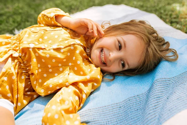 행복한 어린 소녀는 활짝 웃고 잔디에서 담요에 누워 야외에서 여름 날을 즐기고 있습니다. 공원에서 가족과 함께 피크닉을 하는 동안 재미있고 편안한 시간을 보내는 사랑스러운 아이. — 스톡 사진