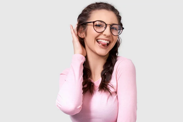 Radosna młoda kobieta pokazująca jej język, migająca z okiem i nosząca okrągłe, modne, przezroczyste okulary i koszulę z długim rękawem, stwarzając na białym ścianie Studio. Pozytywna uśmiechnięta kobieta Making Grimace — Zdjęcie stockowe