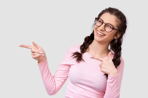 Szczęśliwa młoda kobieta uczeń w przezroczystych okularach, wskazuje na bok z palcami puste miejsce kopiowania dla Ciebie tekst promocyjny, izolowane na białym Studio ścianie. — Zdjęcie stockowe