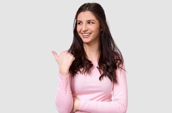 Beautiful European young woman wearing casual pink long sleeve shirt, indicating to blank copy space for advertising text, looking aside and smiling, posing over white studio background. — Stock Photo, Image