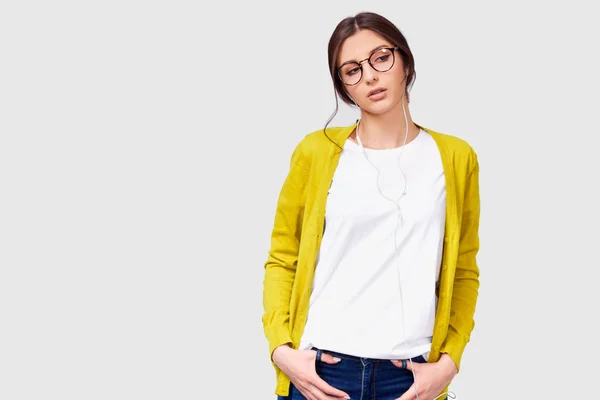 Brunette pretty woman in transparent eyeglasses wearing casual outfit listening to the music from earphones. Indoor shot of Caucasian young female with shiny skin wears yellow blouse and white t-shirt — Stock Photo, Image