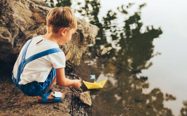 Parkta gölüzerinde sevimli küçük çocuk başlatmak kağıt gemi Yatay arka görünümü. Bir tekne ile oynayan sevimli çocuk çocuk. Çocuk açık havada su kenarında bir oyuncak tekne yelken. Çocukluk ve ekoloji