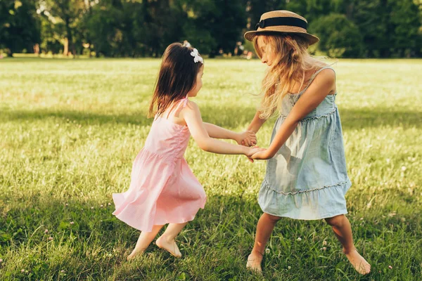 Zwei kleine Mädchen tanzen zusammen auf dem grünen Gras im Park. Kinder spielen und genießen die Sommertage im Park. zwei Schwestern, die sich im Freien an der Sonne amüsieren. Jugend- und Freundschaftskonzept — Stockfoto