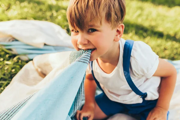 Immagine orizzontale di divertente sciocco ragazzino che gioca con una coperta fuori in giardino. Ragazzo carino divertirsi nel parco durante il picnic con la sua famiglia. Ragazzo allegro che gioca all'aperto — Foto Stock