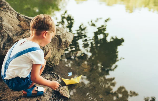 Image de vue arrière de mignon petit garçon lancer bateau de papier sur le lac dans le parc. Adorable gamin jouant avec un bateau. Curieux enfant naviguant sur un bateau jouet au bord de l'eau à l'extérieur. Enfance et écologie — Photo
