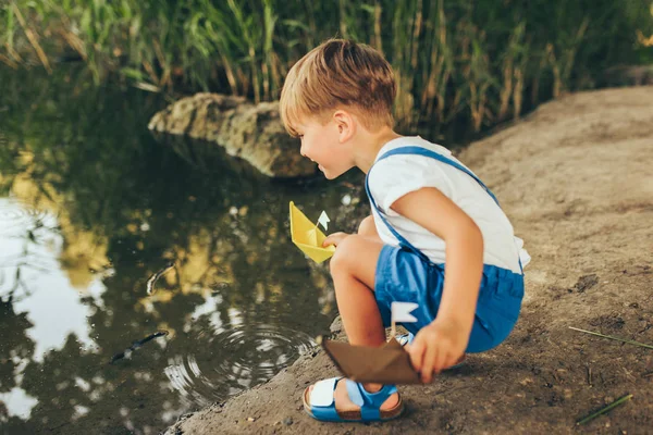 Изображение милого маленького мальчика, запускающего бумажный корабль на озере в парке. Очаровательный мальчик играет с лодкой. Любопытный ребенок, плывущий на игрушечной лодке у воды на открытом воздухе. Концепция детства и экологии — стоковое фото