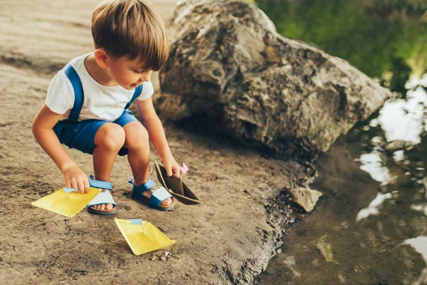 Sevimli küçük çocuk Outdoors görüntü parkta gölüzerinde kağıt gemi başlattı. Bir tekne ile oynayan sevimli çocuk çocuk. Çocuk açık havada su kenarında bir oyuncak tekne yelken. Çocukluk ve ekoloji
