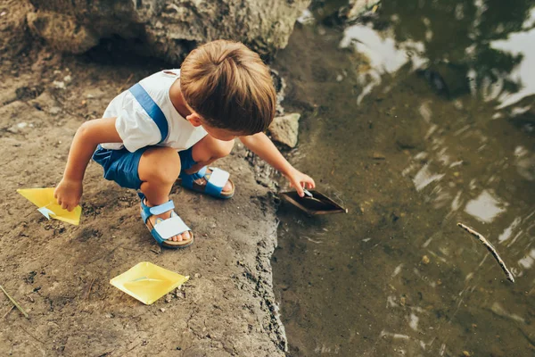 Un bambino adorabile lancia una nave di carta sul lago nel parco. Adorabile ragazzino che gioca con una barca. Bambino che naviga una barca giocattolo dal bordo delle acque all'aperto. Infanzia ed ecologia — Foto Stock