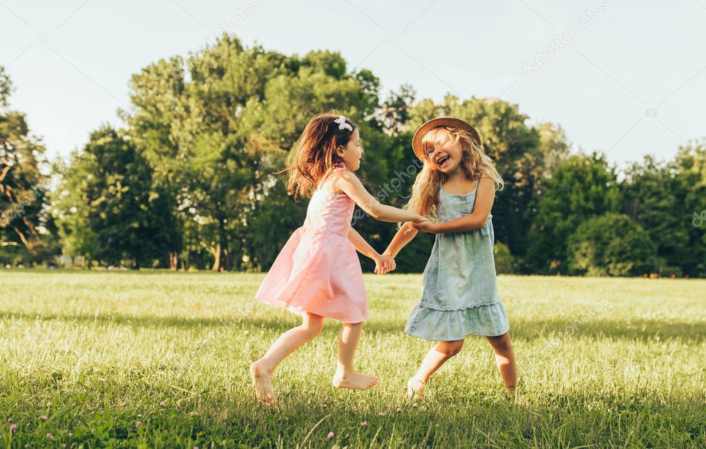 Horizontal image of two little girl dancing on the green grass in the park. Children enjoying summer days in the park. Two sisters having fun on sunlight outdoors. Childhood and friendship concept