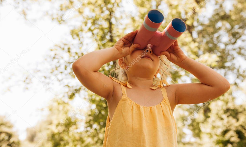 Bottom view image of cute little girl looking through a binoculars searching for an imagination or exploration in summer day in park. Curious child playing pretend safari game outdoors in the forest.