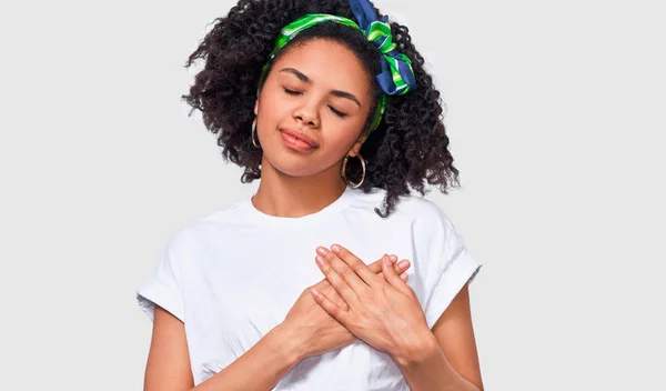 Agradable y agradecida joven afroamericana sonriendo con los ojos cerrados y mantiene ambas palmas en el pecho, vestidas con camiseta blanca, aisladas sobre una pared blanca. Personas, emociones, salud, lenguaje corporal —  Fotos de Stock