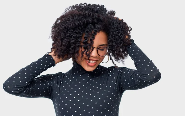 Щаслива темношкірна молода жінка, одягнена в чорний довгий рукав сорочки з білими точками і окуляри, тримаючись руками на її здорове волосся. Афро-американські жінки посміхаючись широкої постановки на білій стіні студії — стокове фото