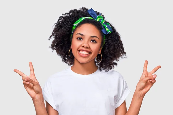 Close-up portret van positieve Afro-Amerikaanse jonge vrouw glimlachend breed, het tonen van vrede gebaar terwijl op zoek naar de camera, staande tegen witte studio achtergrond. — Stockfoto