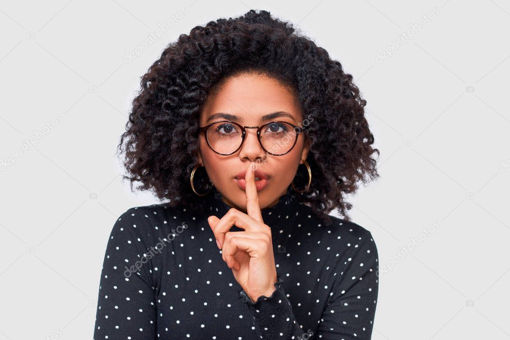 Closeup studio portrait of African American young woman wears black long sleeve shirt, transparent eyewear holding index finger on lips, asking to keep silence. Dark skinned female asks to be quiet.