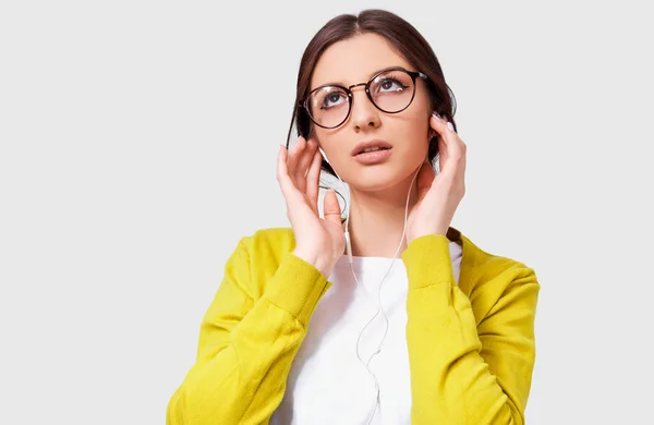 Портрет молодой женщины в прозрачных очках в повседневной одежде, слушающей музыку из наушников. Студийный кадр кавказской студентки, слушающей аудиоуроки на белой стене — стоковое фото