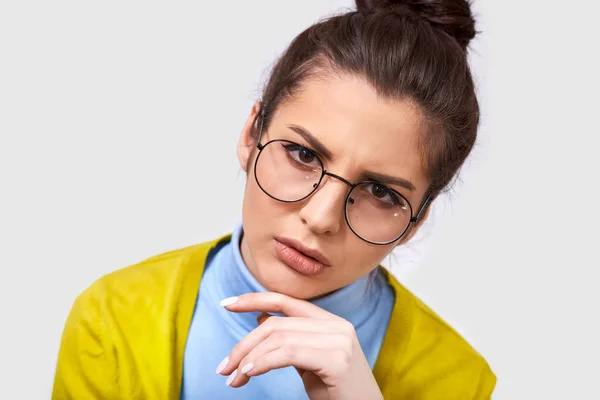 Retrato de cerca de una mujer joven y seria vestida con atuendo casual, gafas redondas transparentes, siendo dudosa, mirando a la cámara. Estudiante europea pensando en exámenes y posando en la pared blanca — Foto de Stock