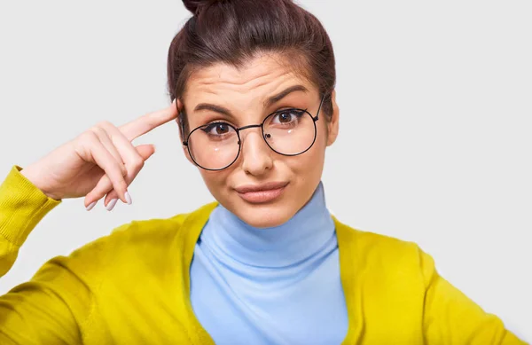 Closeup image of doubtful young woman with a bun hairstyle, wearing casual outfit and round transparent spectacles, looking at the camera, isolated over white studio background. Emotions, confusion — Stock Photo, Image
