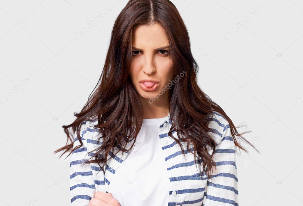 Young woman grimacing and sticking out her tongue having mad feelings, isolated over white background. Pretty student girl wearing casual outfit, having fun and making grimace.