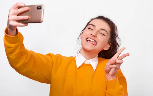 Happy young woman making funny grimace with tongue and peace gesture, wearing orange sweater, taking selfie on smart phone. Pretty girl student making self portrait over white wall. People, emotions — Stock Photo, Image