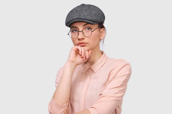Портрет серьезной брюнетки молодой женщины носит прозрачные очки, повседневную рубашку и серую кепку, серьезно смотрит прямо в камеру, позирует на белом студийном фоне. Люди эмоции conce — стоковое фото