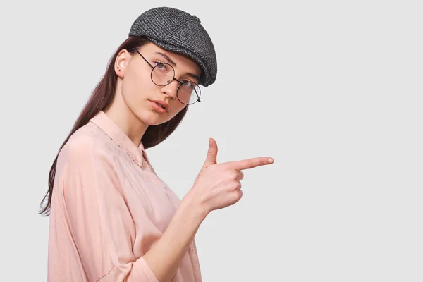 Poziome portret szczery młodej kobiety ubrany różowy shirt i okrągłe przezroczyste okulary, wskazuje z palcem wskazującym pokazuje coś w pustej przestrzeni kopiowania do reklamy, patrząc poważnie. — Zdjęcie stockowe