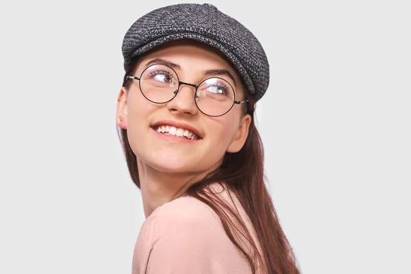 Retrato de close-up da jovem mulher feliz, sorrindo amplamente, usa camisa rosa, boné cinza e óculos redondos transparentes. Estudante bonita se sente alegre e posando sobre a parede do estúdio branco. Pessoas, emoção — Fotografia de Stock
