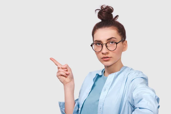 Tiro franco de mujer joven seria lleva camisa azul y gafas redondas transparentes, indica con el dedo índice, demuestra algo en espacio de copia en blanco para la publicidad, mirando a la cámara —  Fotos de Stock