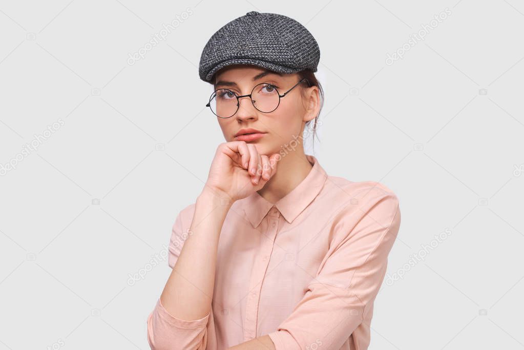 Portrait of serious brunette young woman wears transparent spectacles, casual shirt and gray cap, looks seriously directly into the camera, poses against white studio background. People emotions conce