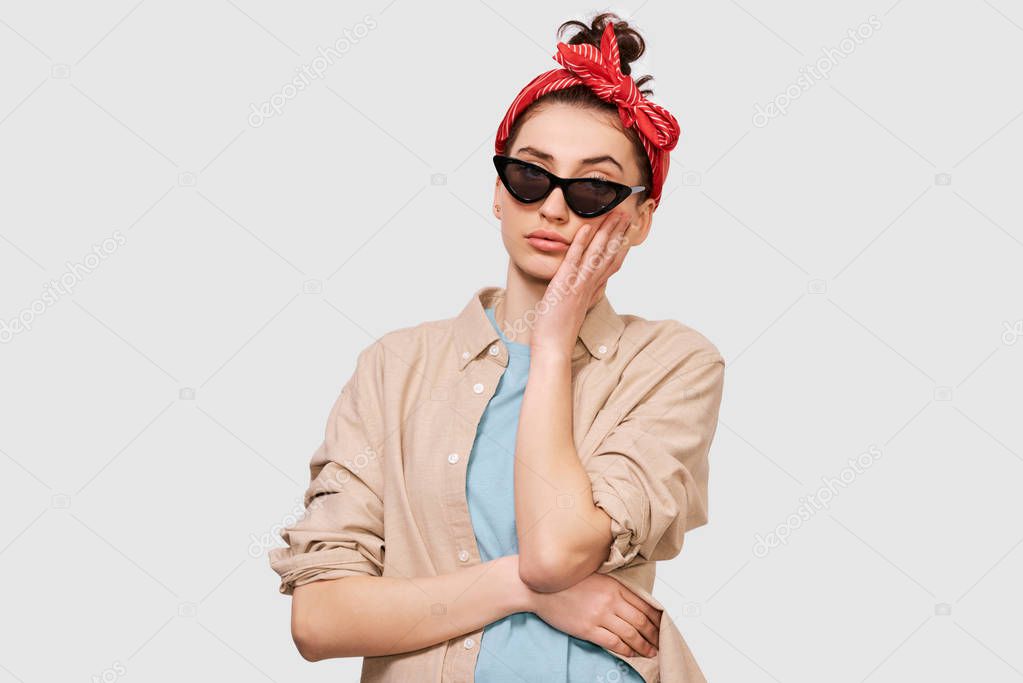 Pensive brunette young woman wears casual outfit and black sunglasses, ooks seriously directly into camera, poses against white studio background. People emotions concept
