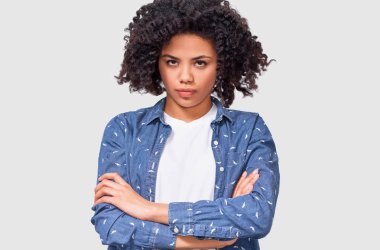 Üzgün genç Afro kadının Görüntü deli yüz ifadesi vardır, rahat kıyafet giyer, elleri çapraz tutar, beyaz stüdyo arka plan üzerinde izole. Üzgün kadın gri duvar üzerinden kameraya bakıyor.