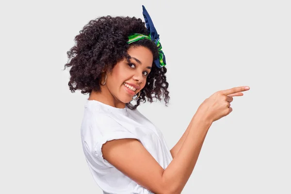 Estudio vista lateral retrato de mujer joven afroamericana vestida con camiseta blanca y diadema verde, indica con el dedo índice a espacio de copia en blanco, aislado sobre la pared blanca . — Foto de Stock