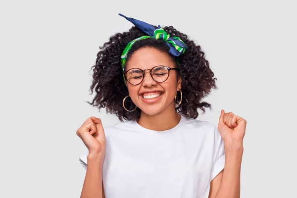 Portrait studio horizontal de jeune femme aux yeux fermés, les poings serrés, se réjouit du succès, porte un t-shirt blanc et un bandeau vert, portant des lunettes rondes transparentes, sur fond blanc — Photo