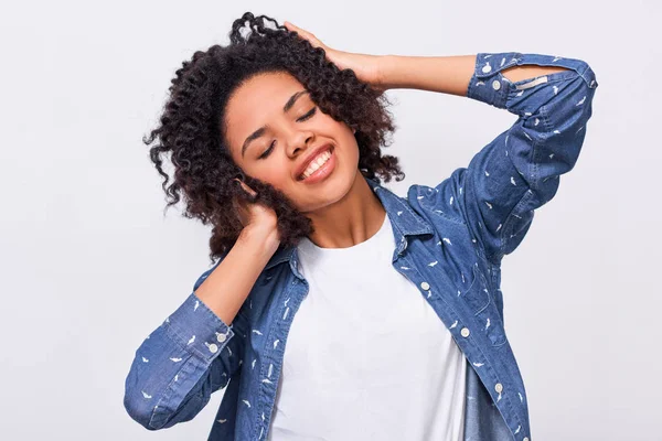 La imagen de la joven de piel oscura y complacida vestida con camisa azul, cogida de la mano en la cabeza, se siente feliz. Mujer afroamericana sonriendo ampliamente y sintiéndose satisfecha, posando sobre una pared blanca — Foto de Stock