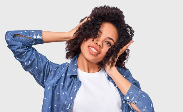 L'image studio d'une jeune femme à la peau foncée, vêtue d'une chemise bleue, tenant la main sur la tête, se sent heureuse. Femme afro-américaine souriante et satisfaite, posant sur un mur blanc — Photo