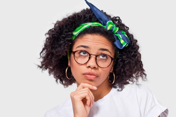 Close-up portret van de kleine Afrikaanse Amerikaanse jonge vrouw die serieus kijkt door transparante brillen op iemand, gekleed in wit t shirt, poses instudio. Student meisje heeft denken expressie — Stockfoto
