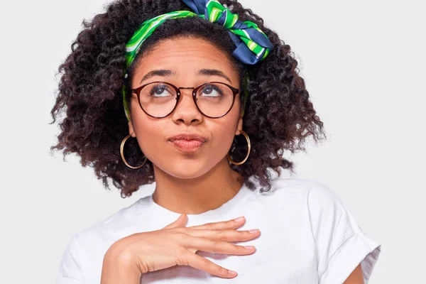 Closeup studio retrato de uma jovem afro-americana pensativa olhando para cima através de óculos transparentes com a mão no peito, vestida com camiseta branca. Estudante menina tem expressão de pensamento — Fotografia de Stock