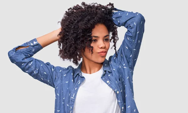 Une jeune femme à la peau foncée vêtue d'une chemise bleue, tenant la main sur la tête, se sent heureuse. Femme afro-américaine souriant largement et se sentant satisfaite, posant sur un mur blanc — Photo