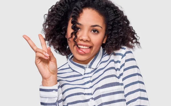Primer plano retrato del estudio de la joven afroamericana positiva sonriendo ampliamente, mostrando gesto de paz y muestra la lengua mientras mira a la cámara, de pie sobre fondo blanco del estudio . —  Fotos de Stock
