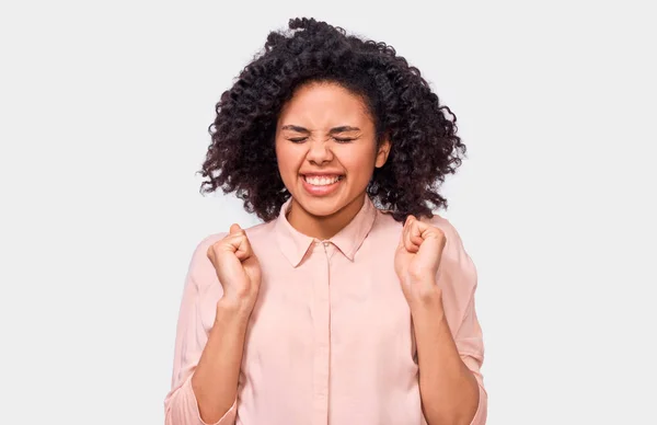 Die Gewinnerin afrikanisch-amerikanische junge Frau ballt mit geschlossenen Augen die Fäuste, trägt ein rosafarbenes Hemd, isoliert vor weißem Hintergrund. Menschen, Erfolg und Emotionen — Stockfoto