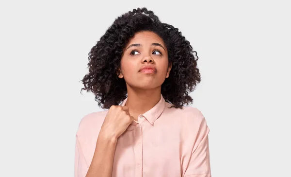 Mujer joven afroamericana disgustada, mirando hacia arriba con expresión dudosa, vestida con una camisa rosa casual, posando sobre una pared blanca. La estudiante tiene expresión confusa. Personas emociones —  Fotos de Stock