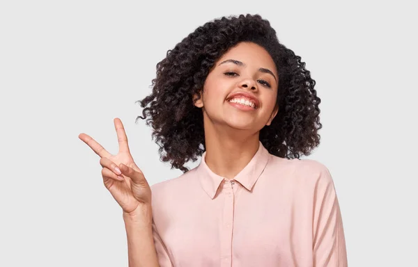 Retrato de estúdio da jovem afro-americana positiva sorrindo amplamente, mostrando gesto de paz enquanto olha para a câmera, de pé contra fundo de estúdio branco . — Fotografia de Stock