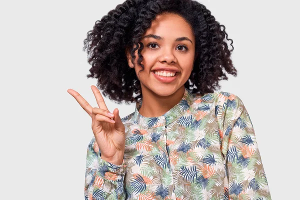 Fechar a imagem horizontal da jovem afro-americana positiva sorrindo amplamente, mostrando gesto de paz enquanto olha para a câmera, de pé contra o fundo do estúdio branco . — Fotografia de Stock